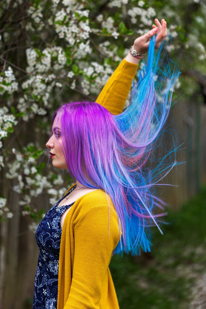 About Woman with purple and blue hair in yellow sweater amidst blooming spring flowers.